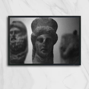 Fotografía análoga en blanco y negro de tres esculturas antiguas de piedra; la figura central, enfocada, muestra un rostro humano con tocado, mientras dos figuras laterales aparecen desenfocadas en primer plano y fondo. Imagen tomada en Udine, Italia, obra Ruinas Udine de Fiorella Angelini, 2018.