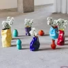 Instalación de Jorge Cabieses-Valdés compuesta por varios jarrones de cerámica esmaltada de formas irregulares y colores intensos —rojo, azul, amarillo, turquesa y morado— dispuestos directamente sobre el piso de una galería. Algunos contienen ramos de crisantemos blancos y se organizan en un conjunto escultórico sobre el suelo de concreto. Cerámica esmaltada, pisos eléctricos y crisantemos, medidas variables, 2023.